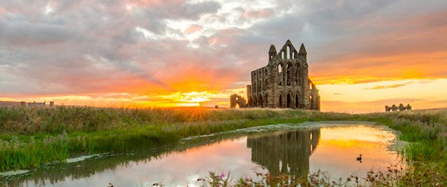 Whitby Abbey viewed from across a river. The sun is rising in the orange sky.
