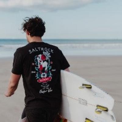 Man in saltrock t shirt walking on beach carrying a surf board